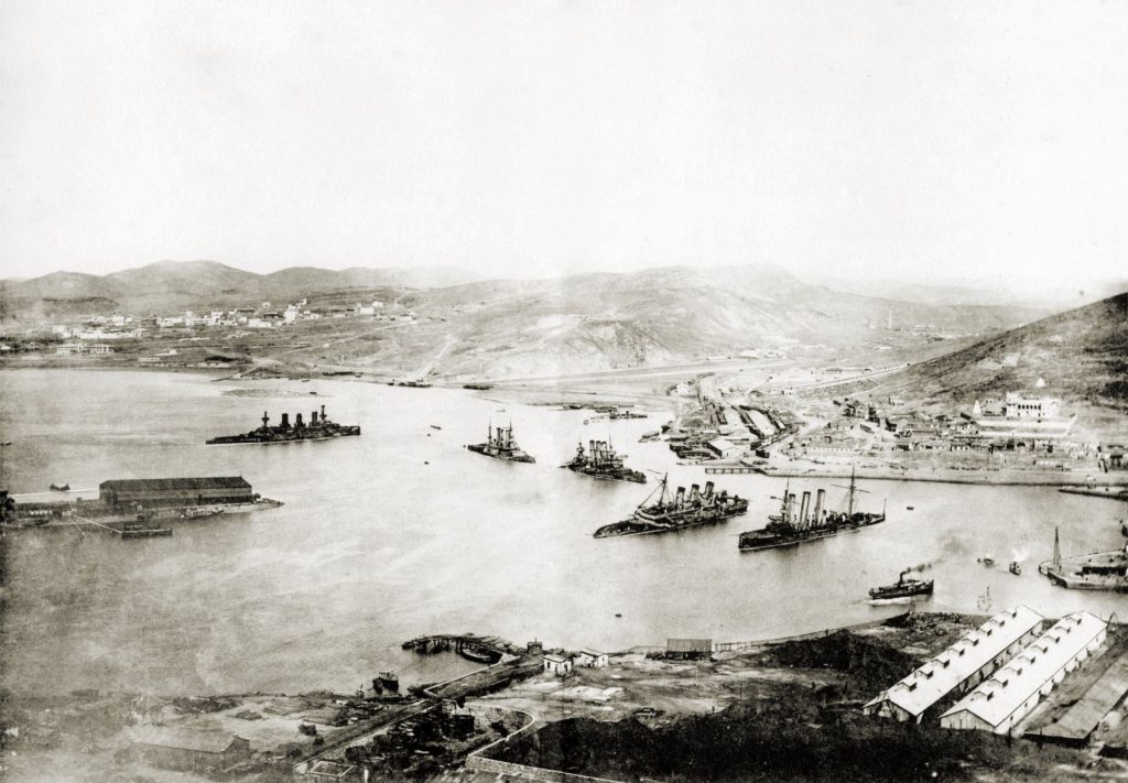 旅順要塞攻略後の旅順港の当時の様子のモノクロ写真。港には何隻か戦艦が浮かんでいる。