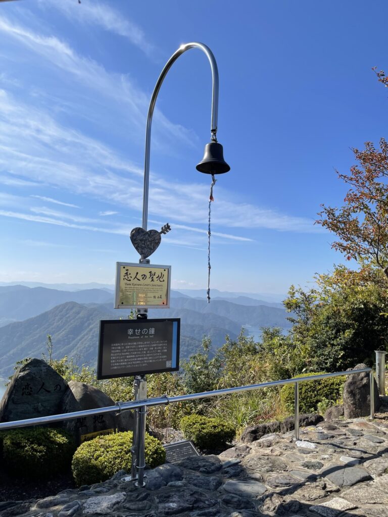 恋人の聖地に置かれた幸せの鐘。晴れた青空の中、背景には壮大な自然と緑の絶景が広がっています。