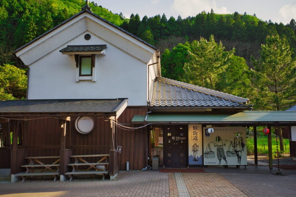 鯖街道にある白壁の2階建て建物。一階部分はこげ茶色の板壁。入り口には付近には、赤と白の提灯や荷物を背負った鯖街道を歩いた昔の人が描かれた看板があります。