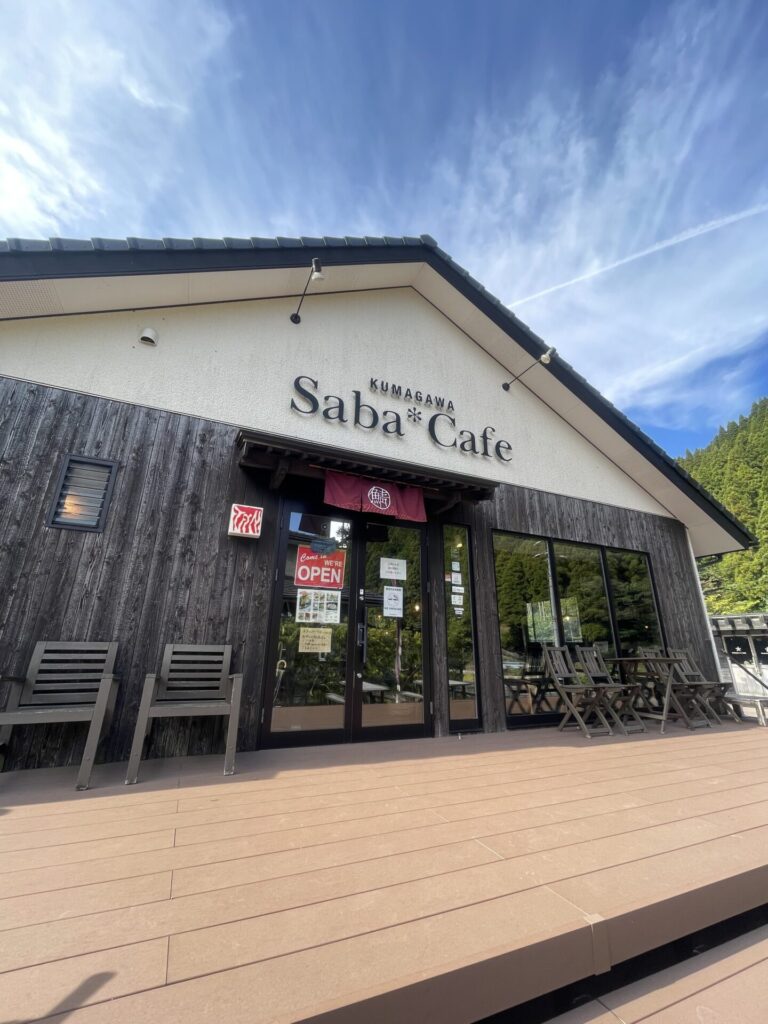 『Saba*cafe』外観。三角屋根の平屋。屋根に近い半分は白色、下はこげ茶色の木材壁で昔ながらの雰囲気があるオシャレな建物。店の入り口にあるウッドデッキには、椅子やテーブルがあります。