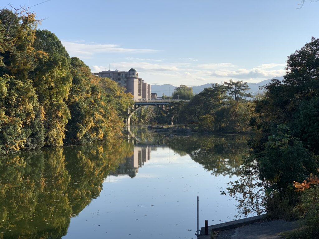 綾川町を流れる綾川。川岸の木々が水面に反射して映り込み、奥には橋と一部の街並みが映っています。