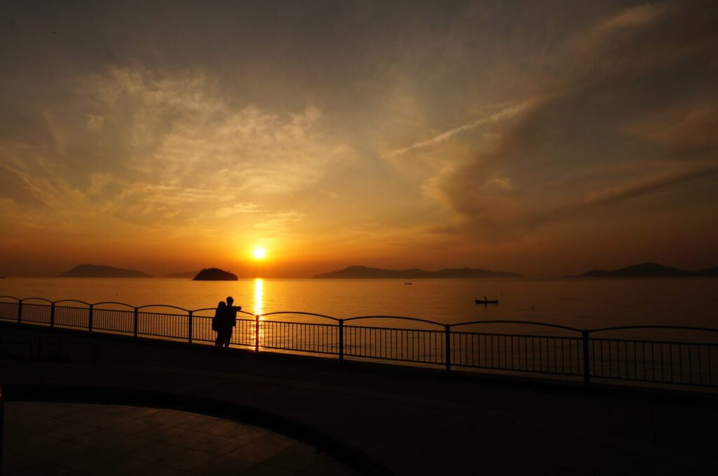 宇多津臨海公園の遊歩道で夕焼けを眺めるカップルが映っています。奥に見える瀬戸内海には夕日が反射して金色に輝いています。