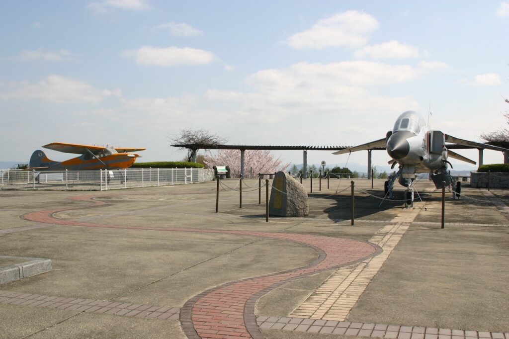 高山航空公園の広場の様子。右手に銀色のジェット機、左手奥にオレンジ色のヘリコプターが映っています。