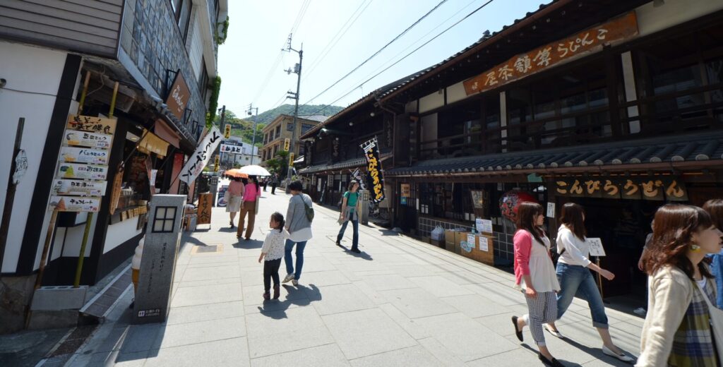 道の両脇には、飲食店やお土産物を販売する店が並ぶ風景。こんぴらうどんやせんべいなどののぼりや看板が見える。人々が行き交っている。