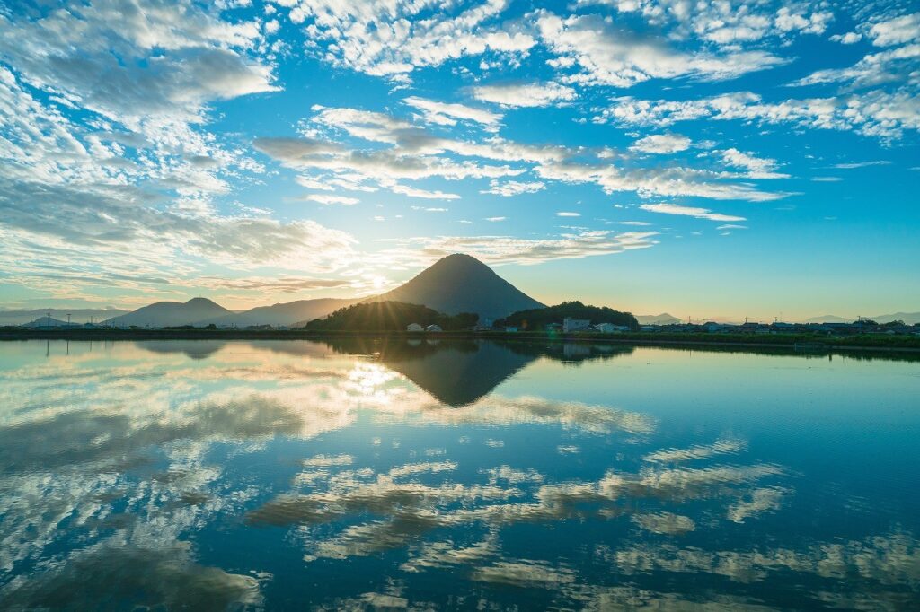 朝日を背景になだらかな裾野が美しい飯野山（通称：讃岐富士）のシルエットが中央に移っており、手前には朝焼けの空と飯野山が水面に反射して映り込んでいます。