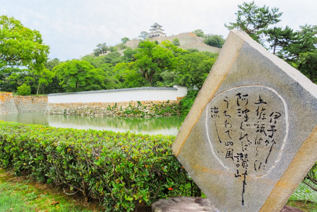 遠くには丸亀城が見え、その周辺には緑の木々が茂っています。堀の前に石碑があり、「伊予竹に土佐紙貼りて阿波で糊付ければ讃岐うちわで四国の涼しい」と刻まれています。