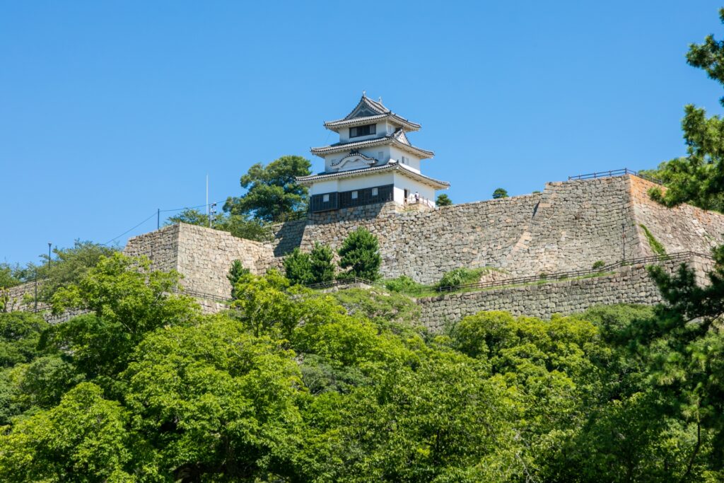 石垣の上に建つ丸亀城。空は澄み渡っており、周囲は緑豊かです。手前には鮮やかな緑の木々が生い茂っています。