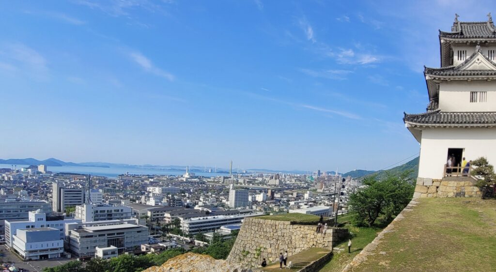 丸亀城の本丸からの景観。画像右端に天守閣があり、その背後に広がる丸亀市と瀬戸内海の広大な景色が晴れの空のもと映えている。