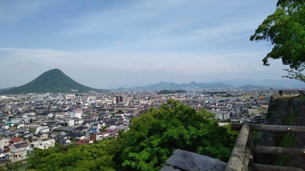 丸亀城の本丸から望む丸亀市の風景。前景には石垣が見え、中景には市街が広がっています。背景には緑豊かな山々が連なり、町の中心にはなだらかな裾野が美しい讃岐富士が見えます。