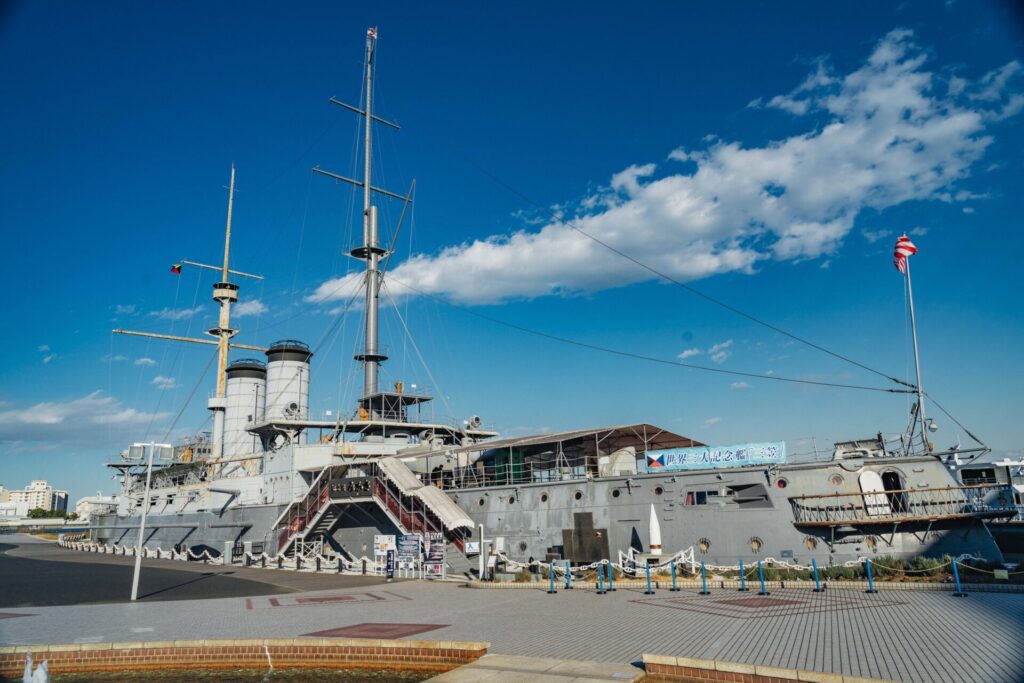 晴れた青空の下、重厚な船体の軍艦「三笠」が写されています。艦橋、煙突、マストが見えます。船の前には、広場が広がっています。