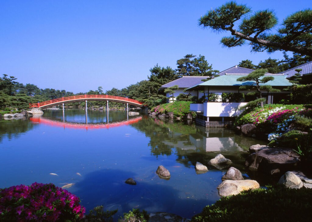 青空の下、穏やかな雰囲気の中津万象園の風景。鮮やかな朱色の橋が静かな池の上を優雅にアーチを描き、手入れが行き届いた松や木々が水辺を囲み、咲き誇るピンクと白の花が緑豊かな風景に色彩を添えています。