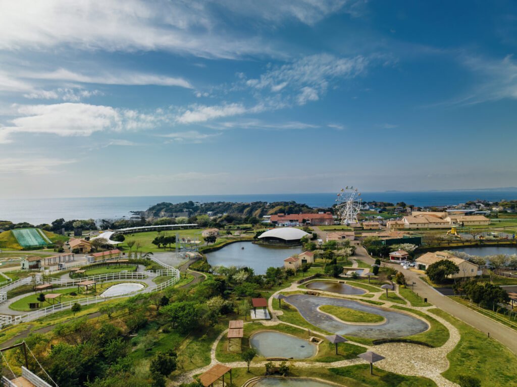 長井海の手公園ソレイユの丘のパノラマビュー。美しい青空と海が背景に広がり、園内には観覧車を含むアトラクション、緑の草地、そして大小さまざまな湖が見えます。