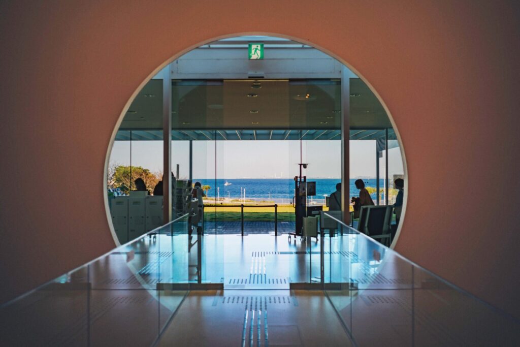 横須賀美術館の内部からの風景。丸いアーチ型の開口部とガラス製のドアを通して、その先には青い海と空が広がっています。中には人々がいます。
