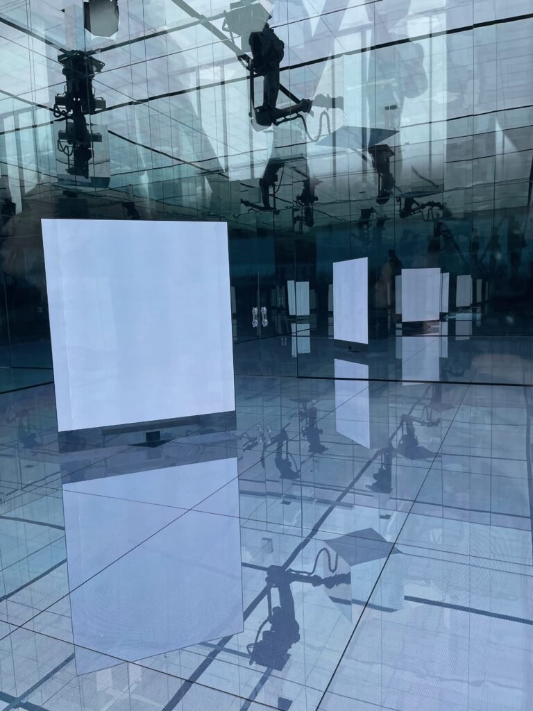 大阪・関西万博の落合陽一プロデュース「null²（ヌルヌル）」パビリオンの内部。青白い光の中、湾曲した鏡面壁が無限の合わせ鏡を作り出し、LEDの床と来場者の影がどこまでも続く幻想的な没入空間。