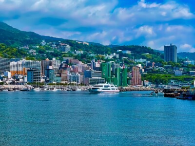 緑豊かな山の麓に広がる熱海港の景色。海には複数の白いフェリーやボートが停泊しており、背後には高層ビルや中層の建物が立ち並んでいる。建物の間には緑の木々が点在し、丘の上まで住宅が続いている。