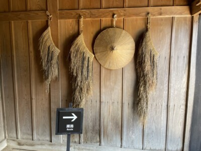 箱根関所内の木造の壁に掛けられた伝統的なわら製の道具や笠。壁には「順路」と書かれた案内標識が設置されており、見学者に進む方向を示している。木の温もりが感じられる内装で、歴史的な雰囲気が漂っている。