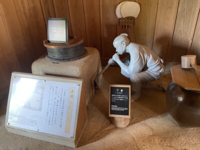 箱根関所内の展示物。白い人形がかがんで吹き矢を吹いている様子を再現している。周囲には木製の壁と伝統的な道具が配置されており、土間の説明パネルが立てられている。展示物には「手を触れないでください」との注意書きも見える。歴史的な関所の生活や仕事の様子を伝える展示がなされている。