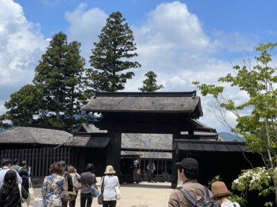 箱根関所の正門に向かって歩く観光客たち。門は黒い木造の伝統的な建築様式で、背後には背の高い木々がそびえ立っている。観光客はカジュアルな服装で、手にカメラやバッグを持っている。背景には青空と白い雲が広がり、歴史的な雰囲気と自然の美しさが調和している。