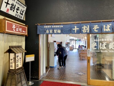箱根関所 旅物語館内のそば処「十割そば」の入口。入口上部には「十割そば 自家製麺」と書かれた青いのれんが掛かっており、左側には「そば処」の看板が見える。店内にはテーブルと椅子が並び、数名の客が食事を楽しんでいる様子が見える。木製の看板と和風の内装が特徴で、落ち着いた雰囲気が漂っている。