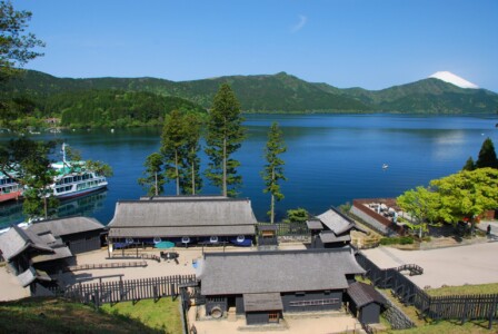 青い湖と緑豊かな山々を背景に、箱根関所跡港が広がっている。湖には遊覧船が停泊しており、右奥には雪をかぶった富士山が見える。手前には歴史的な建物が並び、伝統的な日本の関所の雰囲気を再現している。木々が立ち並び、穏やかな自然と歴史的な景観が調和している。