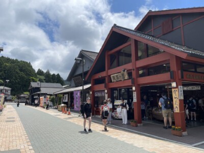 箱根関所通りの賑やかな風景。伝統的な木造建築の建物が並び、その中には「民芸の里」と書かれた木の看板が掲げられているお店がある。観光客が通りを歩きながら店を見て回っており、お土産店や飲食店が並んでいる。左側には緑豊かな木々が広がり、青空には白い雲が浮かんでいる。和の雰囲気が漂う観光地の様子が伝わる。