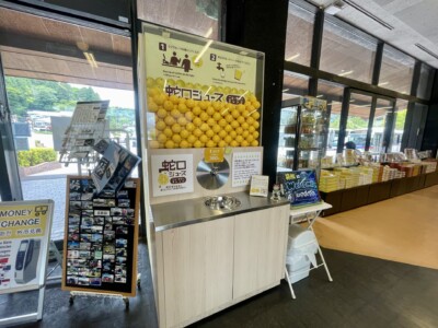 箱根関所 旅物語館内の一角。蛇口から伊豆産のニューサマーオレンジジュースを注げる「蛇口ジュース」のコーナーがあり、上部にはたくさんのニューサマーオレンジのディスプレイがある。ジュースの注ぎ方を説明する看板が掲げられており、その隣には様々なお土産や商品が並んでいる棚が見える。外の景色もガラス越しに見えており、館内は明るく開放的な雰囲気が漂っている。