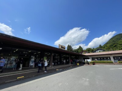 箱根関所 旅物語館の外観。建物には「旅物語館」と書かれた看板が掲げられている。観光客が建物の前で立ち話をしたり、館内に入っていく様子が見える。背景には緑豊かな山々と青空が広がり、雲が浮かんでいる。和風の建築が特徴的で、観光地としての賑わいが感じられる