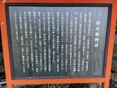 「初木姫物語」と題された木製の案内板。板には白い文字で、初島の守護神である初木神社と初木姫についての物語が詳細に書かれている。案内板の周囲は赤い枠で囲まれており、背景には石や木の根が見える。