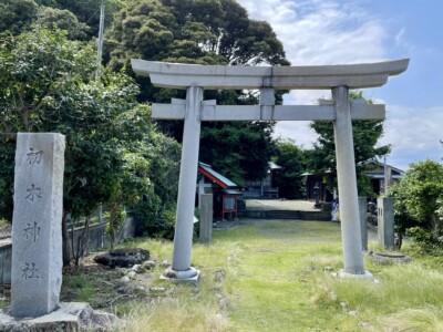 「初木神社」の鳥居と石碑が立つ参道の入り口。鳥居の背後には緑豊かな木々が茂り、神社の建物が見える。鳥居の右側には「初木神社」と刻まれた石碑が建っており、参道は草が生い茂っている。神社の静寂な雰囲気が漂っている。
