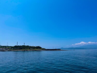 静かな海と緑豊かな初島の半島が見える風景。海岸線には低い建物と数本の通信塔が立っている。遠くには山々が連なり、その上には白い雲が浮かんでいる。穏やかな水面と広がる青空が印象的な景色である。