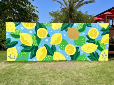 色鮮やかなレモンと葉のデザインが描かれたウォールアート。切られたレモンや丸ごとのレモン、そして緑の葉が散りばめられており、背景は青空を思わせる水色。左下には「hatsushima」の文字が青で書かれている。緑の芝生の上に設置されており、ヤシの木や赤いパラソルの下で楽しむリゾート地の雰囲気が漂っている。