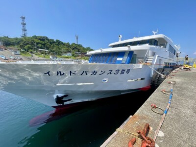 港に停泊している「イルドバカンス3世号」という名前の白いフェリー。船はロープで岸壁に固定されており、船首には黒いアンカーが見える。背景には緑の木々と建物、そして通信塔が立っている丘がある。青い空と澄んだ水面が広がり、快晴の下でのんびりとした雰囲気が漂う。