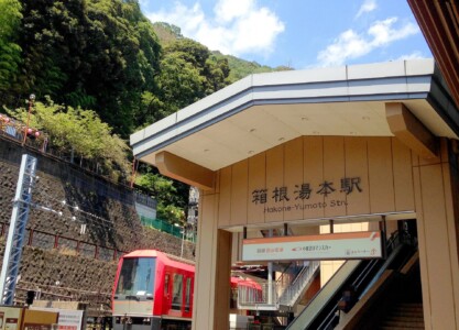 箱根湯本駅の入口。駅名が「箱根湯本駅」と書かれた看板が見える。建物の背後には緑豊かな山々が広がり、青空が広がっている。駅のプラットフォームには赤い電車が停車している。右側にはエレベーターの案内も見える。