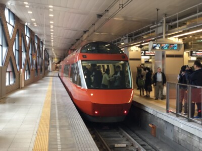 駅のホームに停車中の赤い小田急ロマンスカー。ホームには乗客や見送りの人々がいて、電車の車内にも乗客が見える。背景には「特急ロマンスカー」と表示された看板やホームの案内板がある。