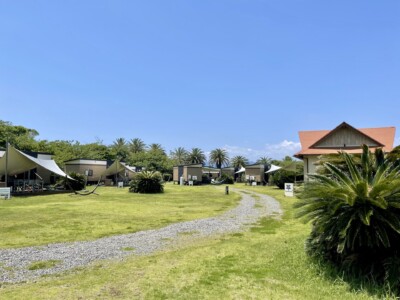 広々とした芝生のエリアに立ち並ぶ小さなコテージやテントが見えるリゾート地の風景。左側にはハンモックが設置されたテントがあり、右側には赤い屋根の建物が見える。背景にはヤシの木が点在し、リゾートの雰囲気を醸し出している。中央には砂利の道が通り、静かでリラックスした雰囲気が漂っている。全体的に、自然に囲まれた快適な宿泊施設が提供されている。