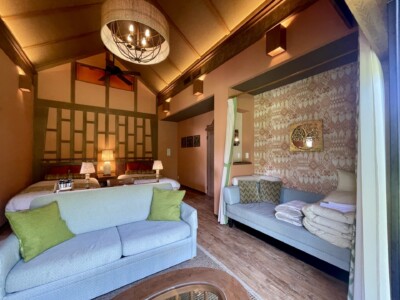 温かみのあるリゾート宿泊施設「アジアンリゾート ヴィラ」の一室。部屋には2つのベッドがあり、ベッドサイドにはランプが置かれている。中央にはライトグリーンのクッションが置かれたソファがあり、その向かいには木製のテーブルが見える。右側には装飾が施された壁とカーテンで仕切られたスペースがあり、そこにはもう一つのソファと追加の寝具が置かれている。天井にはシャンデリアが吊るされ、全体的に落ち着いた雰囲気が漂う。