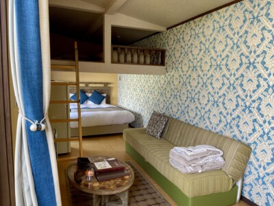 明るい色調で装飾されたリゾート宿泊施設の一室。壁には青と白の柄模様の壁紙が貼られ、シンプルで落ち着いたデザインのソファが置かれている。ソファにはクッションとたたまれたタオルケットが置かれている。手前には木製のテーブルがあり、その上には書類や小物が置かれている。奥には大きなベッドがあり、青いクッションがアクセントになっている。ベッドの上にはロフトスペースがあり、はしごでアクセスできるようになっている。カーテンも青と白のデザインで、部屋全体に統一感がある。