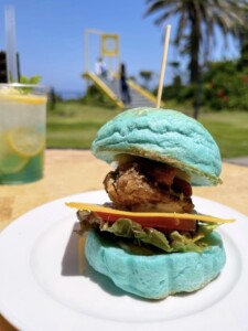 鮮やかな青いバンズのハンバーガーが白い皿に盛り付けられている。ハンバーガーにはフライドチキン、チーズ、レタス、トマトが挟まれており、上から竹串が刺さっている。背景にはレモネードのカップと、黄色いドアのオブジェが見える。リゾート地の庭園で提供されるユニークな食事が、自然の美しい景色と共に楽しめる様子がうかがえる。