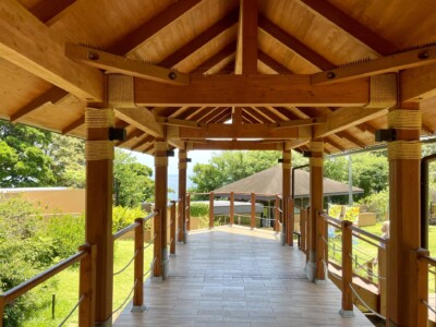 木製の屋根付き廊下の内部。梁や柱には縄が巻かれ、細部にこだわった作りが見える。両側にはロープの手すりが設置されており、外には緑豊かな風景が広がっている。奥には別の建物の屋根と、明るい青空が見えている。
