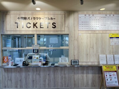 「十国峠パノラマケーブルカー」のチケット売り場の写真です。木目調の壁に「TICKETS」と大きく書かれた看板があります。窓口には透明なガラスがあり、各種案内やポスターが貼られています。右側には時刻表と料金表が掲示されています。