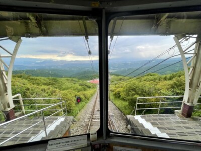 ケーブルカーの車内から撮影された景色が写っています。窓越しに見えるのは、緑豊かな山々と遠くに広がる平野の景色です。レールは急な斜面に沿って下り、左右には手すりと階段があります。遠くには市街地や雲の間から差し込む光が見え、広大な自然が広がっています。