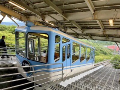 駅のホームに停車している水色と白色のケーブルカーが写っています。ケーブルカーの前部には「十国峠」と書かれた紺色の標識があります。ケーブルカーの周囲には手すりがあり、乗客が乗り降りするための階段が見えます。背景には緑豊かな景色が広がっており、遠くに赤い屋根の建物も見えます。ホームの上部には金属製の屋根があり、駅全体が覆われています。