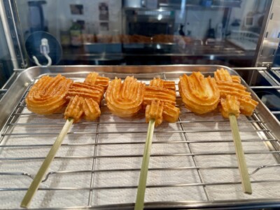 ショーケースの中に並べられた十国峠名物の「峠チュロス」が写っています。「峠」の文字をかたどったチュロスは竹串に刺さっており、サクサクとした食感が感じられる焼き色がついています。チュロスはショーケース内の金網の上に置かれ、キッチンペーパーが敷かれています。背景にはカフェの内部がぼんやりと見え、調理器具やその他の備品が配置されているのが分かります。全体的に、チュロスは美味しそうで、カフェの一品として魅力的に展示されています。