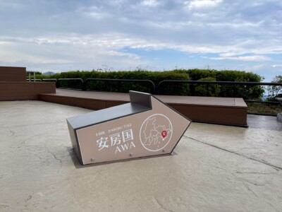 安房国の矢印モニュメント。灰色の矢印型モニュメントが地面に設置されており、「十国峠 JUKKOKU TOGE 安房国 AWA」と書かれています。背景には緑豊かな植生と曇り空が広がっています。矢印の中には地図と位置を示す赤いポイントが描かれています。