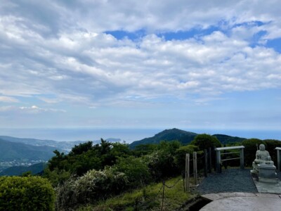 安房国方面の風景。緑豊かな山々が広がり、その先には町と海が見えます。空には雲が広がり、所々に青空が覗いています。写真の右手前には石像があり、周囲にフェンスが設置されています。