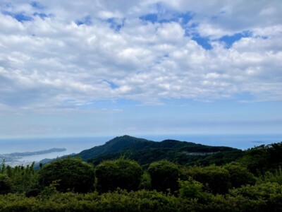 上総国方面の風景。緑豊かな山々が広がり、その先には町と海が見えます。空には雲が広がり、所々に青空が覗いています。