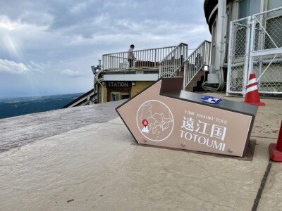 遠江国の矢印モニュメント。灰色の矢印型モニュメントが地面に設置されており、「十国峠 JUKKOKU TOGE 遠江国 TOTOUMI」と書かれています。背景には展望台の一部が見え、手すりに寄りかかって景色を眺める人がいます。矢印の中には地図と位置を示す赤いポイントが描かれています。