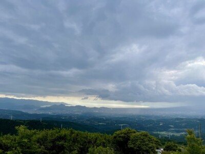 遠江国方面の風景。前景に濃い緑の木々が広がり、背景には山々とその麓に広がる町が見えます。空は厚い雲に覆われており、所々に光が差し込んでいます。