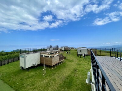 広々とした草原の上に、小さなキャビン風のコテージが並んでいる様子が写っています。建物の一部には屋上テラスがあり、観光客が景色を楽しんでいます。建物は木材と白いパネルで構成されており、周囲の自然と調和しています。背景には青い空と白い雲が広がり、遠くには山々が見えます。手前にはフェンスが設置されており、緑の草地が広がっています。数名の人々が草地に立っており、リラックスした雰囲気が感じられます。