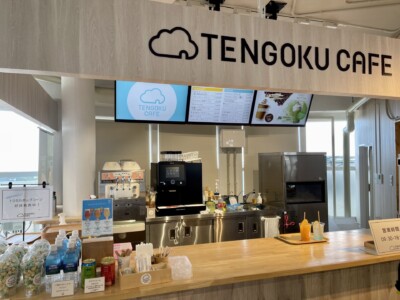 「TENGOKU CAFE」のカウンターが写っています。カフェのロゴが掲げられた看板の下には、コーヒーマシンやソフトクリームマシンが並んでいます。カウンターにはカラフルなドリンクがトレイに乗せられて置かれており、手前にはポップコーンやその他の軽食などがディスプレイされています。上部にはデジタルスクリーンがあり、メニューや商品情報が表示されています。全体的に明るくモダンな内装です。